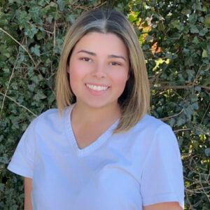 Young woman wearing neutral colored scrubs in front of shrubbery