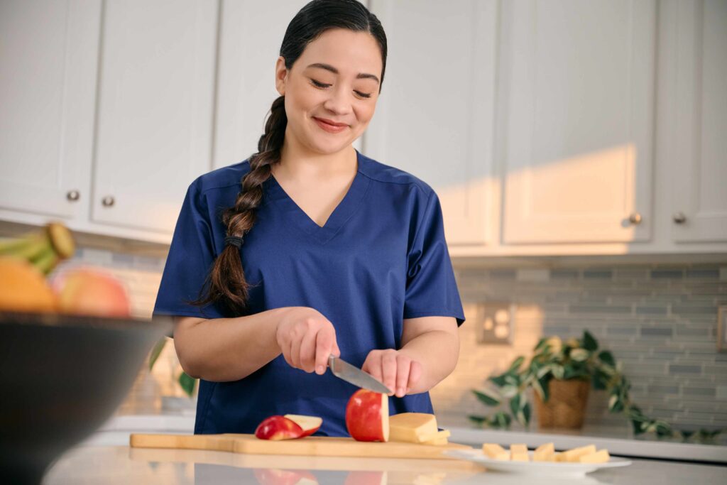 A person in blue scrubs slices an apple in a kitchen with white cabinets and a gray backsplash.