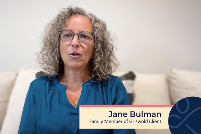 A woman with curly gray hair and glasses sits on a couch, speaking. The lower third of the image reads, "Jane Bulman, Family Member of Griswold Client." She discusses the benefits of local home care services.