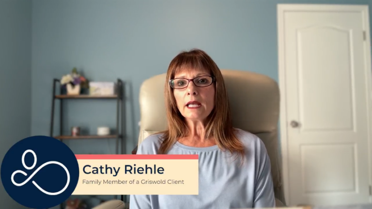 A person with glasses and shoulder-length hair sits in an office chair against a blue wall. A name graphic reads: "Cathy Riehle, Family Member of a Griswold Client.