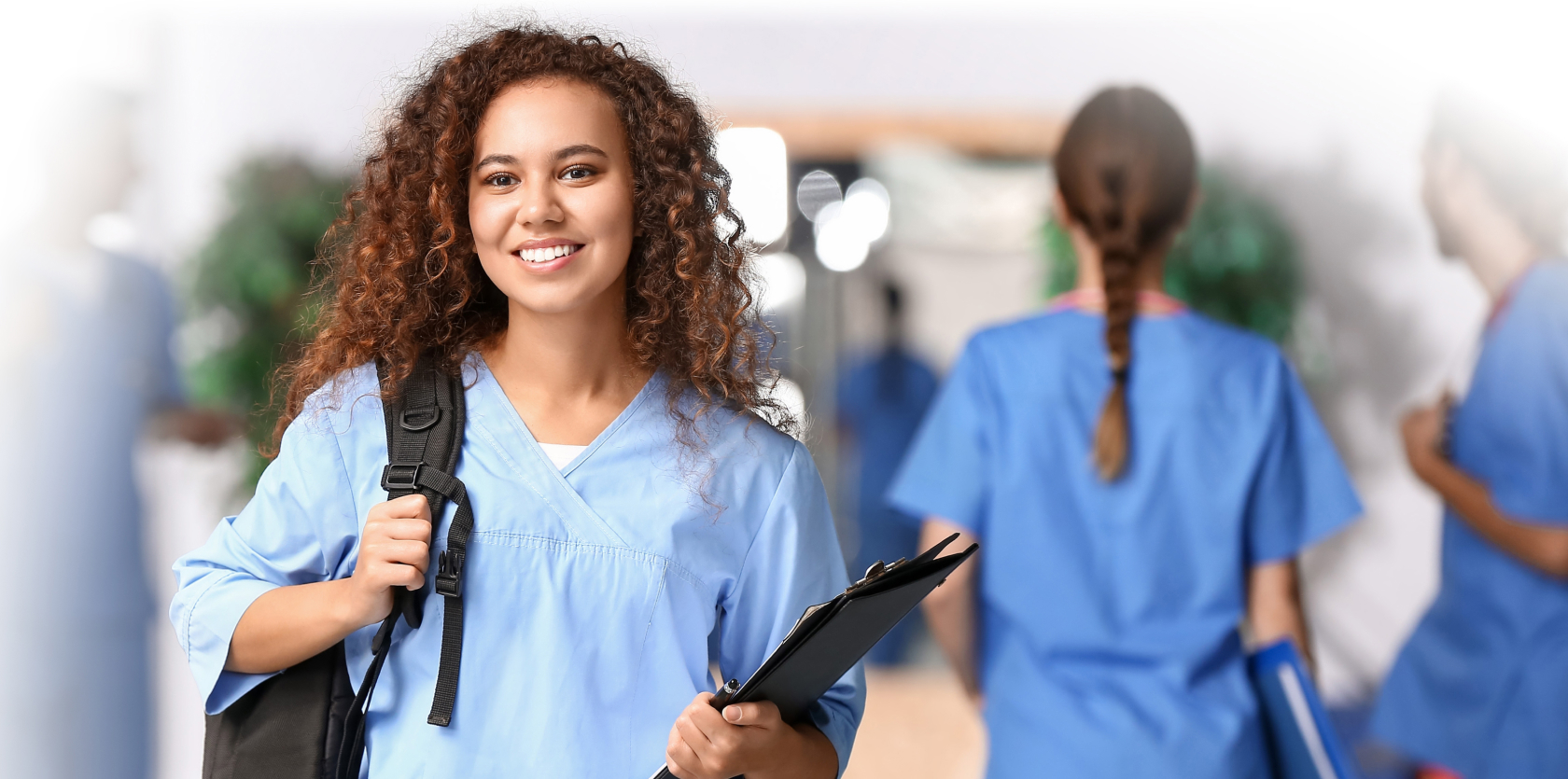 A smiling woman wearing medical scrubs and carrying a backpack and clipboard stands in a hallway, representing personal care services, with two other people in scrubs visible in the background.