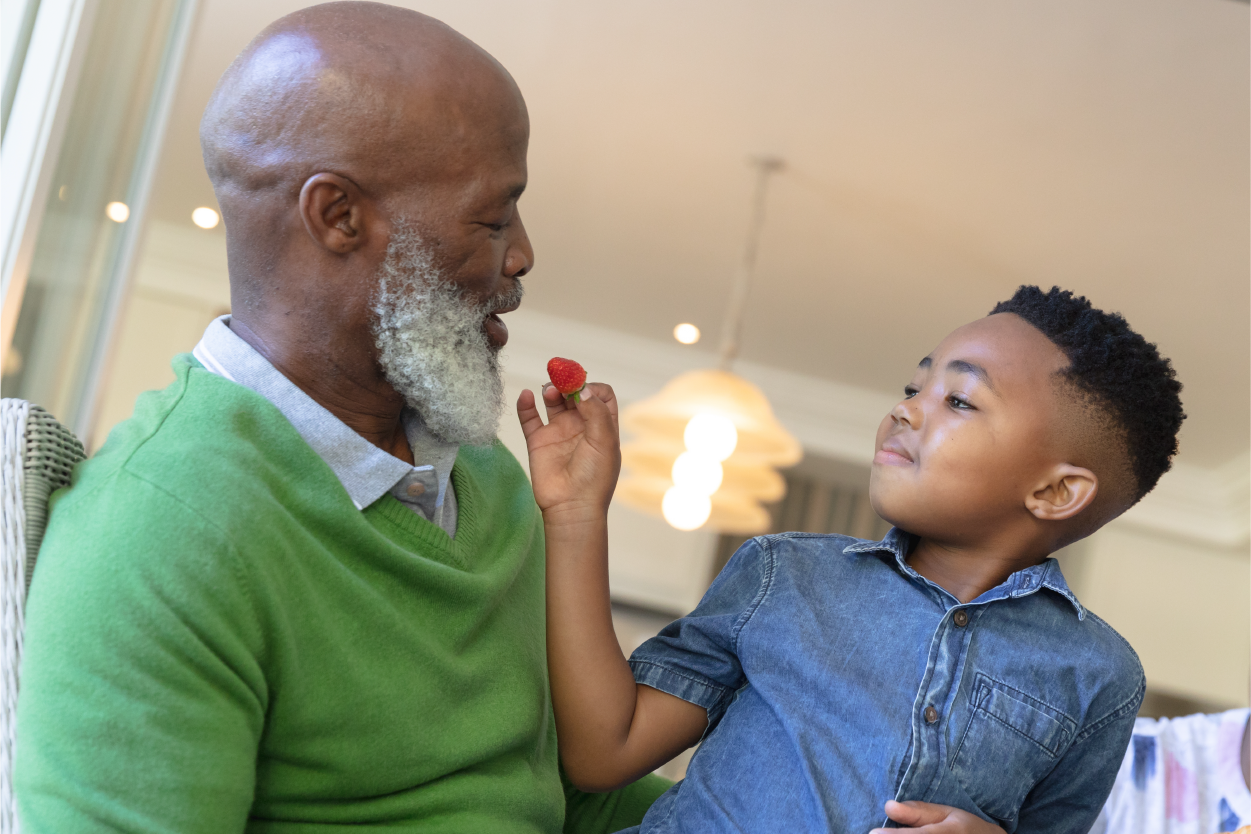 An elderly man with a gray beard smiles at a young boy holding a raspberry towards his mouth