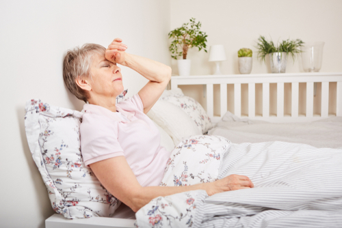 An older woman sits in bed, holding her forehead with a pained expression