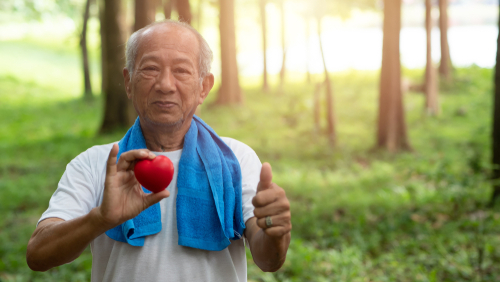 An elderly man outdoors holds a red heart-shaped object in one hand and gives a thumbs-up with the other