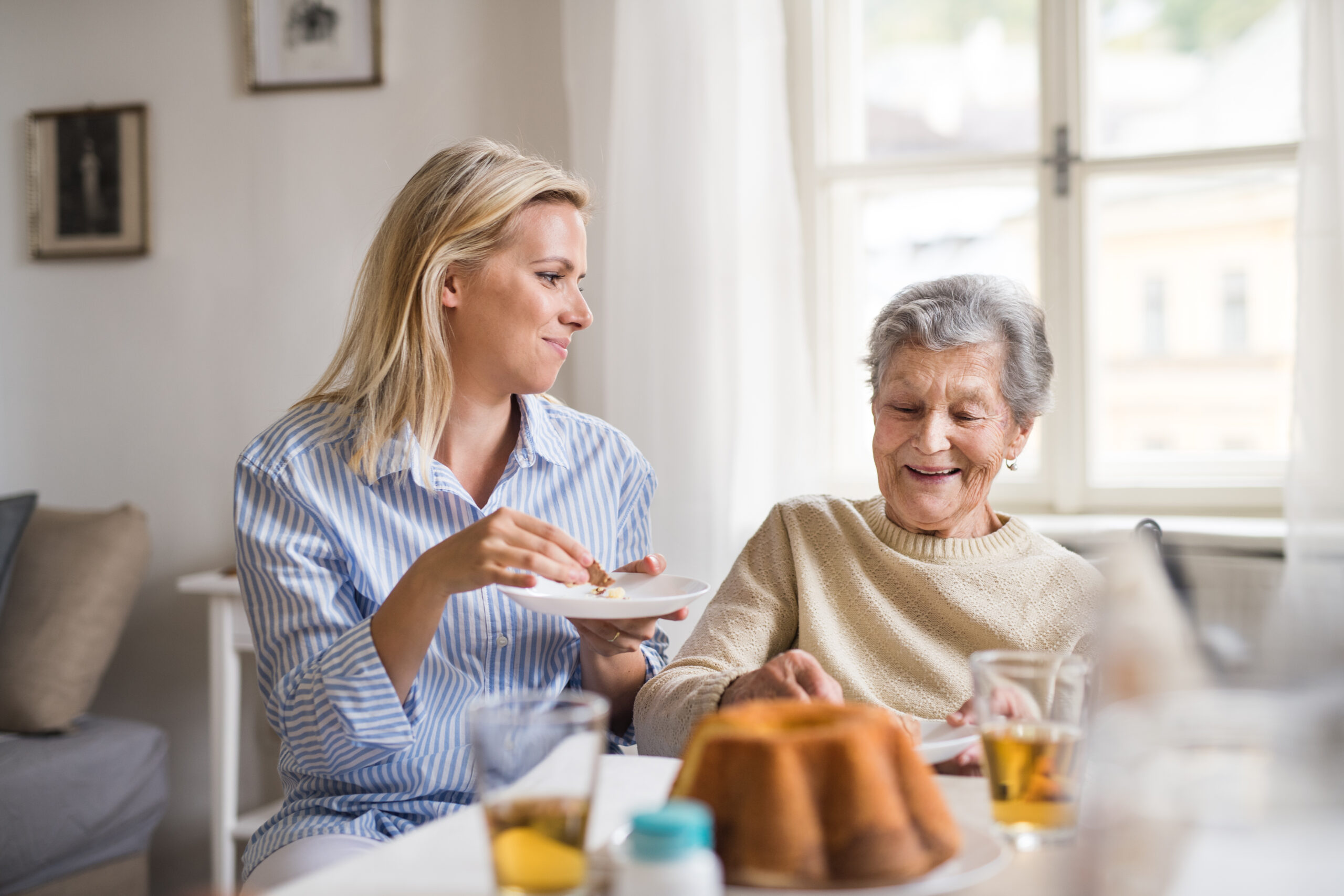 A young woman offers food from a plate to an elderly woman at a dining table.