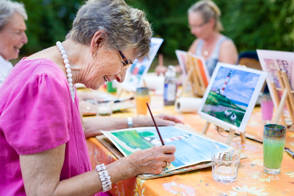 An elderly woman in a pink top is painting a landscape on a canvas at an outdoor table