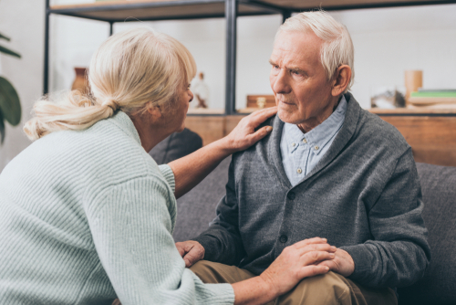 An elderly woman comforts an elderly man sitting on a couch