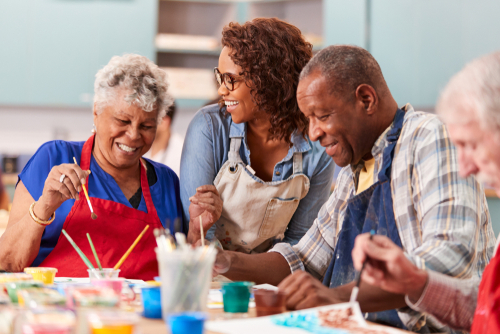 A woman, providing caregiver services, assists two seniors and another person as they paint at a table in an art class