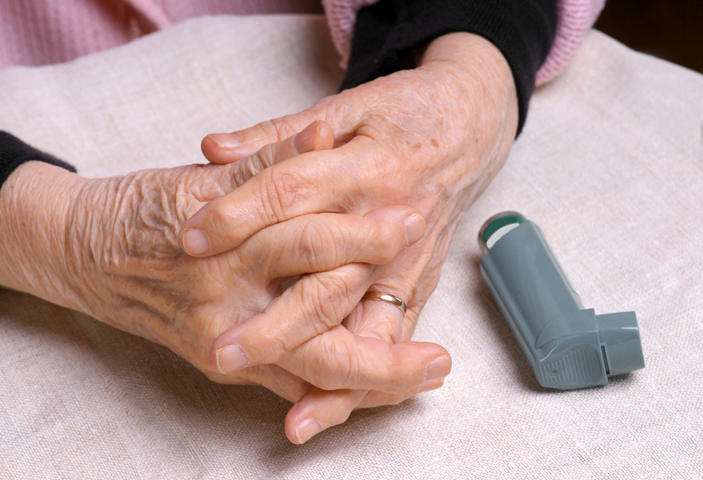 Close-up of elderly hands clasped together on a fabric surface next to an inhaler