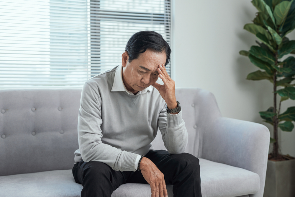 An elderly man sits on a couch with one hand on his forehead