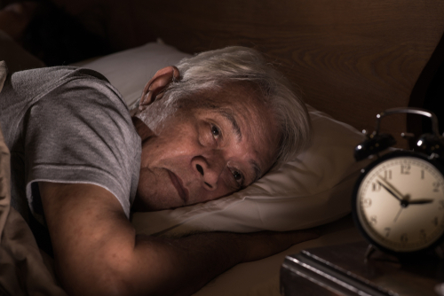 An elderly man lies in bed, awake and looking at an analog alarm clock on a bedside table