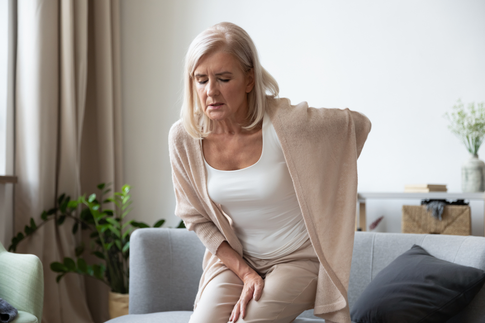 A mature woman sits on a couch, holding her lower back in apparent discomfort