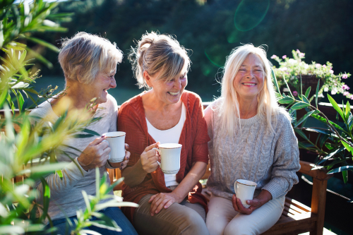 Three women sitting on a bench outdoors, smiling and holding coffee mugs