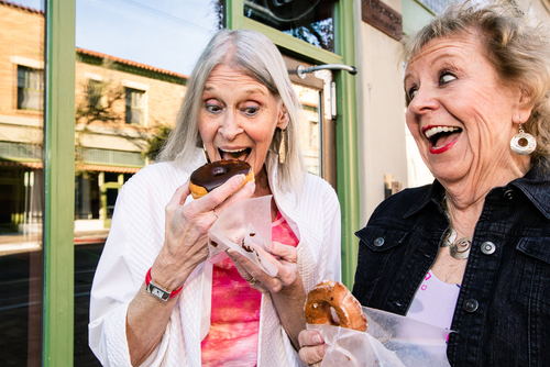 Two women standing outside a storefront, holding and excitedly preparing to eat donuts