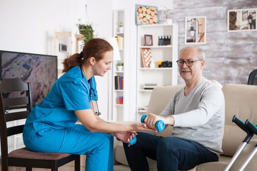 A care giver assists a seated elderly man with arm exercises using a small dumbbell in a living room setting