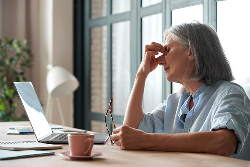 A woman with gray hair sits at a desk holding her glasses in one hand