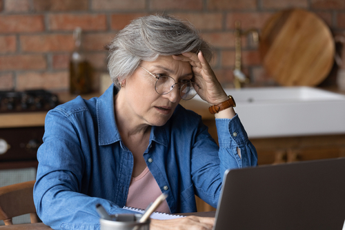 An older woman wearing glasses looks stressed as she engages with a laptop in a kitchen setting