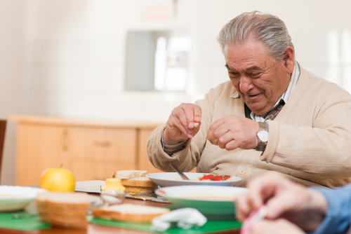 An elderly man is seated at a table, seasoning his meal