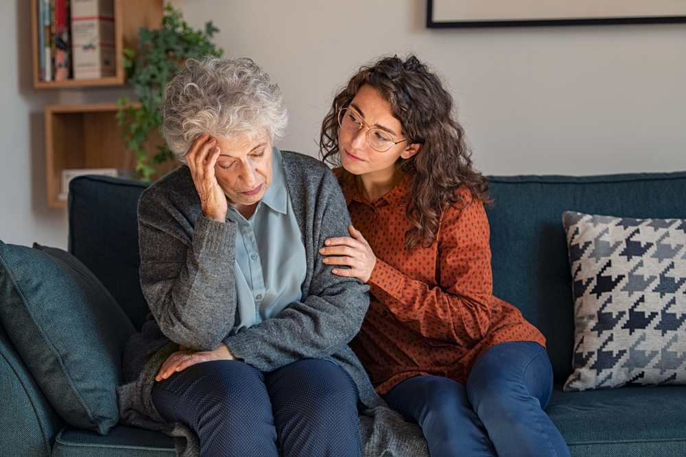 An elderly woman sits on a couch, holding her head in one hand