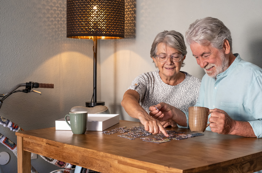 An elderly couple sits at a wooden table, assembling a jigsaw puzzle