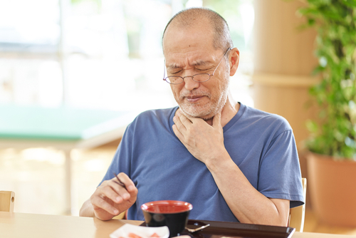 An older man in a blue shirt sits at a table, holding his throat with a pained expression