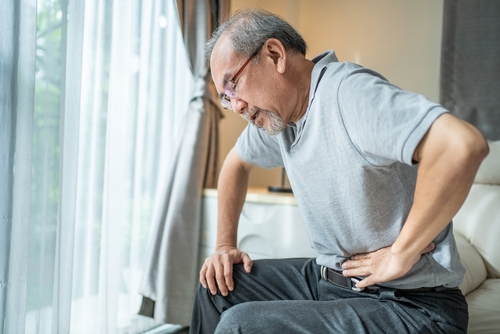 An elderly sits on a couch by a window, holding his side and appearing to be in pain