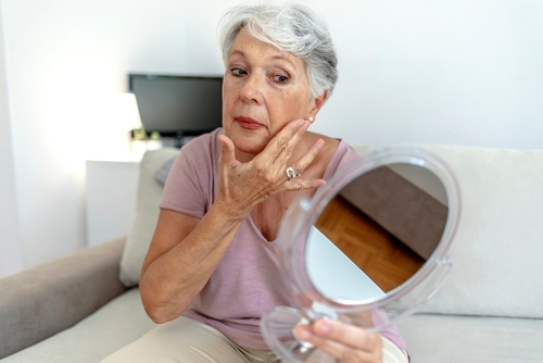 An elderly woman examines her face in a handheld mirror