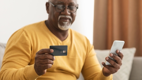 A person wearing glasses and a yellow sweater holds a credit card in one hand and a smartphone in the other while sitting on a couch