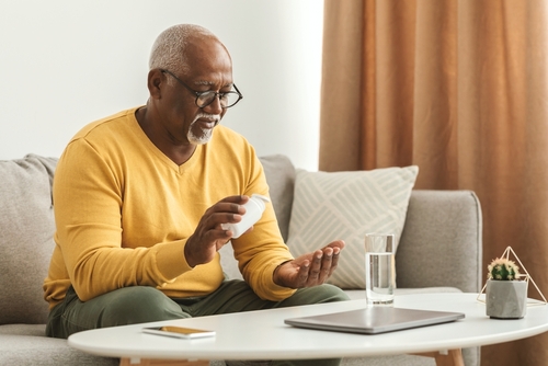 An elderly man wearing glasses sits on a sofa, holding a pill bottle and pouring medication into his hand