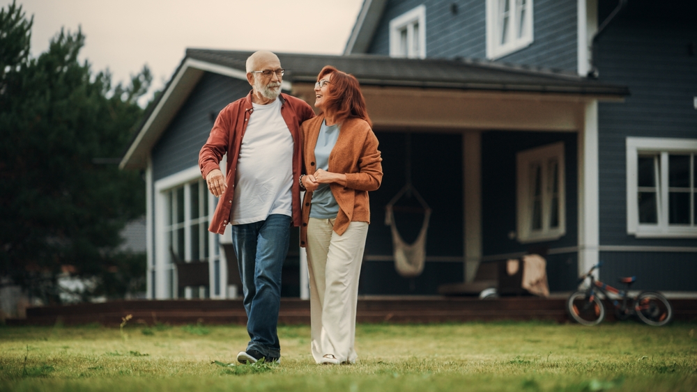 An older couple walks arm in arm on the grass in front of a blue house