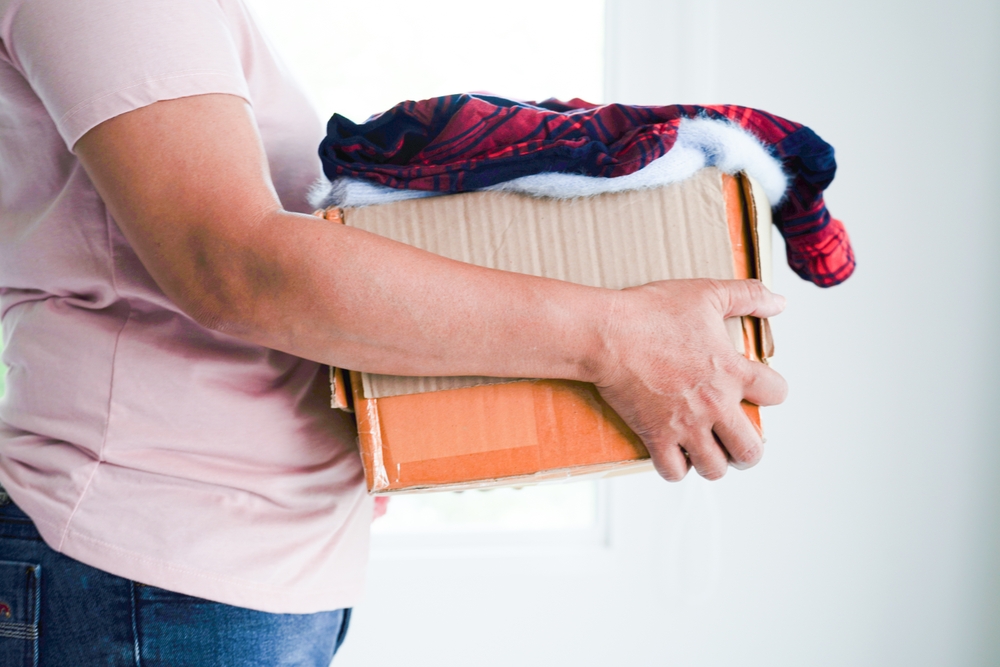 A person holds a cardboard box with clothes spilling out