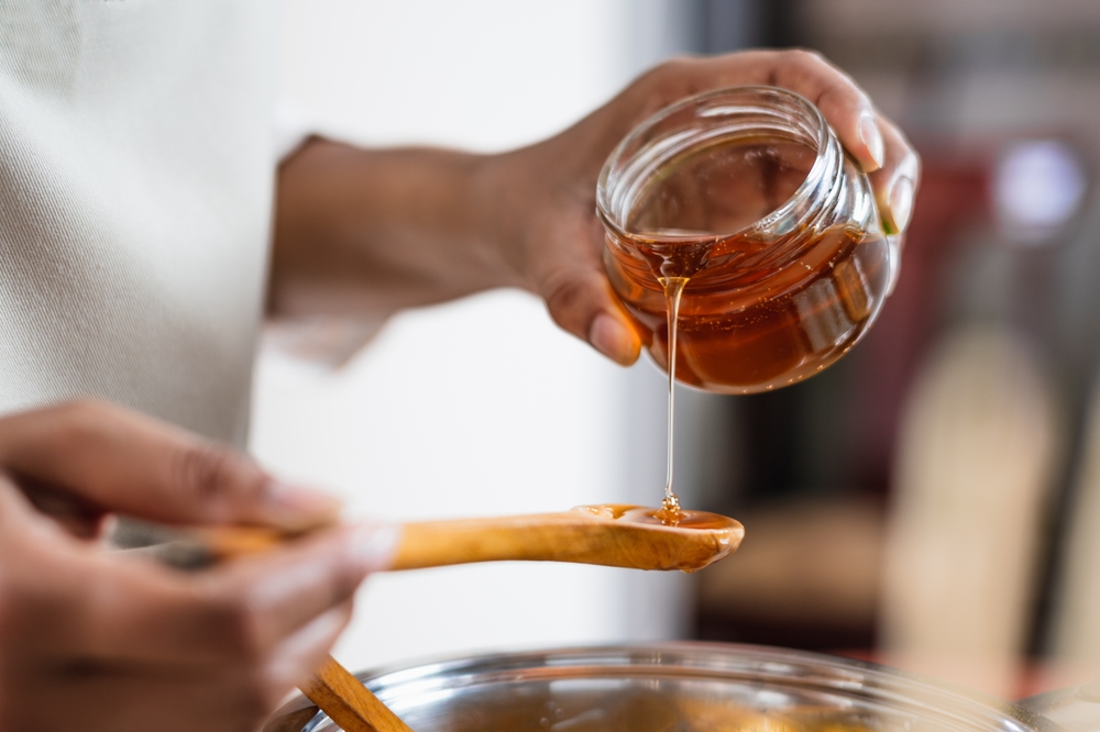 A person pours honey from a glass jar into a wooden spoon