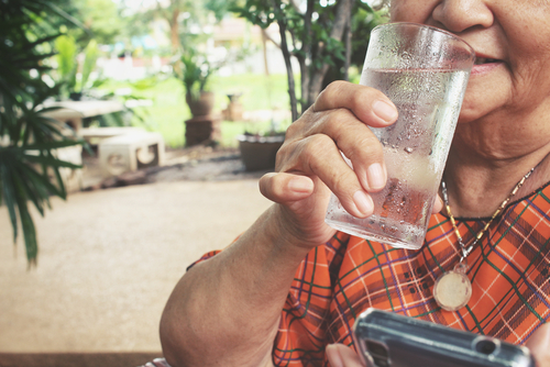 An elderly person drinks water from a glass while holding a smartphone