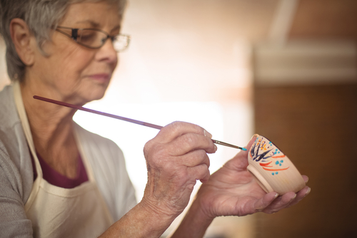 An elderly person wearing an apron carefully paints a small ceramic cup with a fine brush