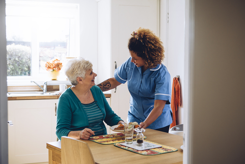 A caregiver in a blue uniform smiles and assists an elderly woman seated at a table with food