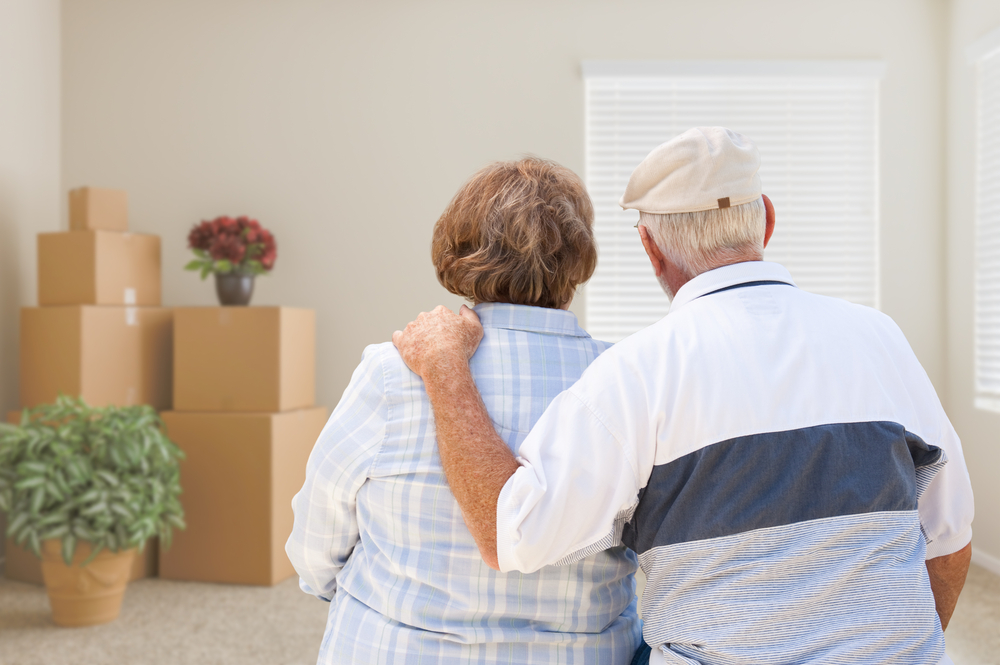 An elderly couple stands closely together, facing a room with packed moving boxes