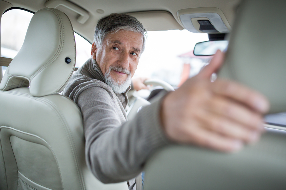 An elderly man looks over his shoulder while sitting in the driver's seat of a car