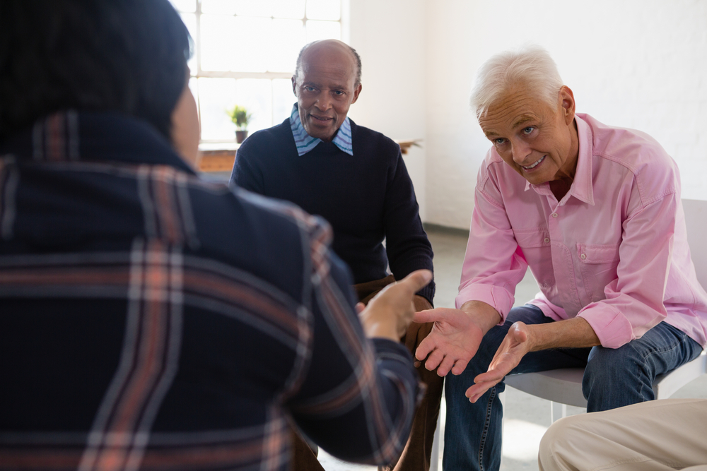 Two elderly men sit and converse with a third person