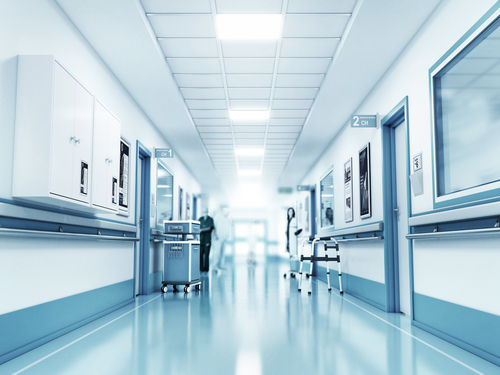 A bright, clean hospital corridor with medical equipment, wall cabinets, and a nurse standing in the background