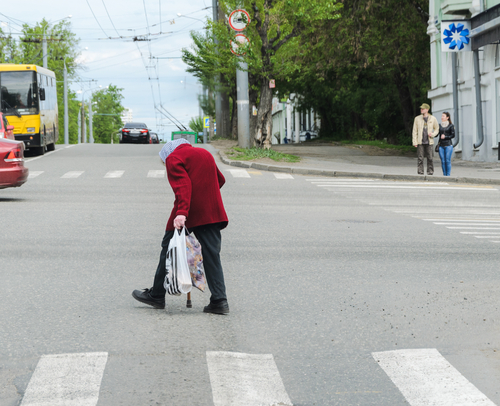 An elderly person with a cane and a shopping bag crosses a street