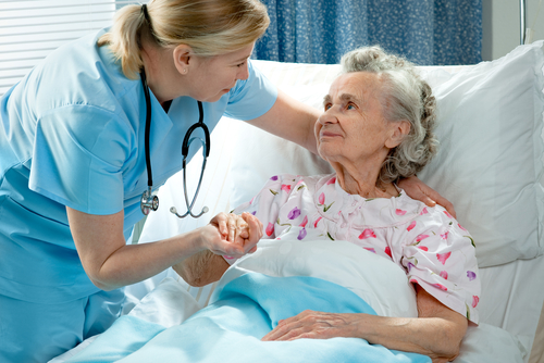 A nurse in blue scrubs holds the hand of an elderly woman lying in a hospital bed