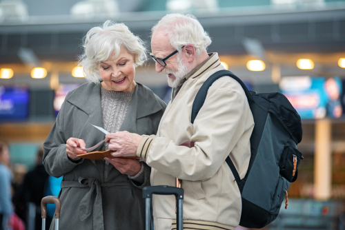 Two elderly people, stand together at an airport, looking at travel documents.