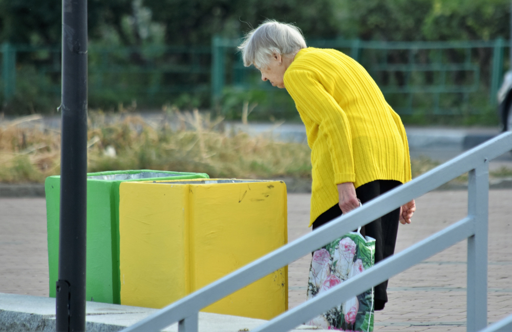 An elderly person in a yellow sweater stands next to a set of colorful recycling bins