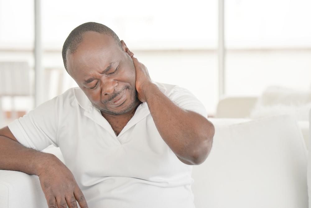 A man in a white shirt sits on a couch, holding his neck with a pained expression