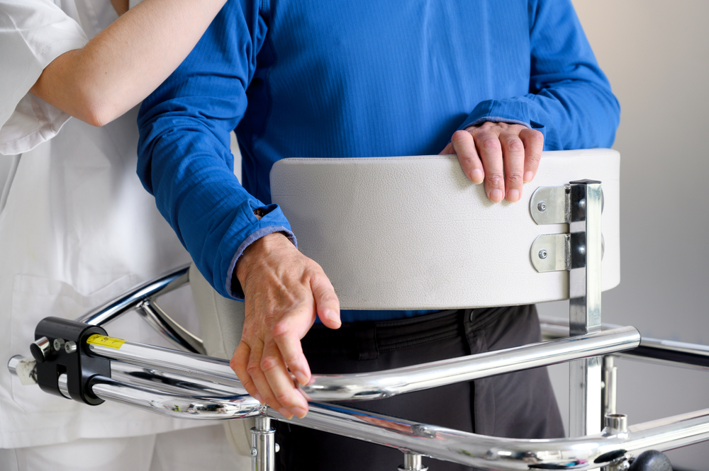 A medical professional from a home health care service assists a patient wearing a blue shirt using a mobility aid.