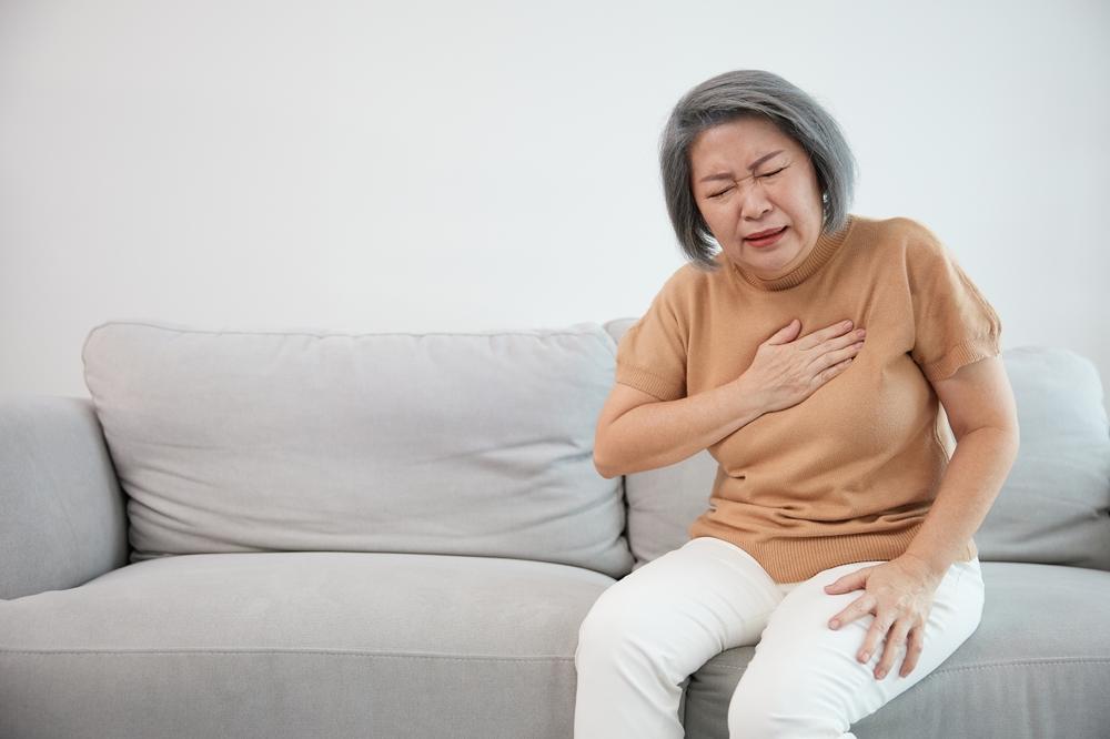 An older woman sits on a sofa, clutching her chest with a pained expression