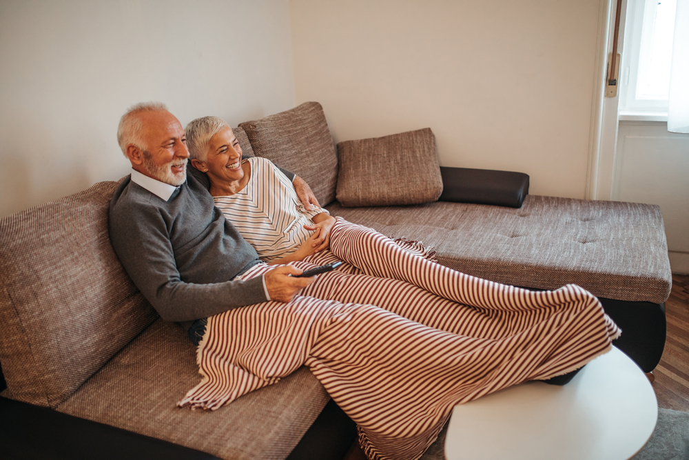 An elderly couple sits on a couch, smiling and covered with a striped blanket, while watching TV