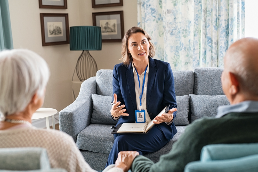 A professional caregiver in a suit sits on a couch, engaging in a conversation with an elderly couple