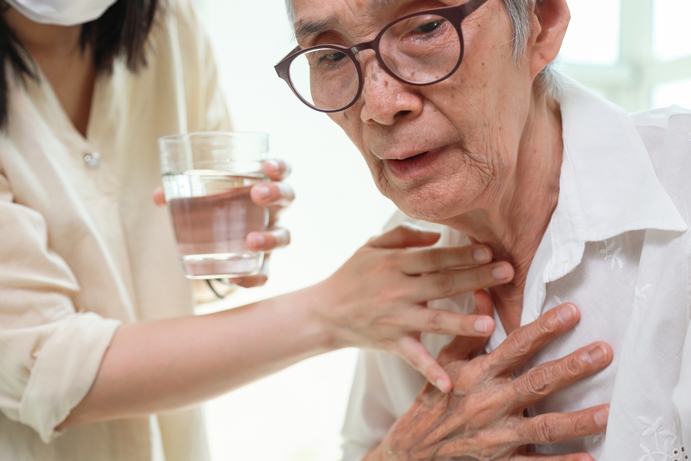 An elderly person wearing glasses holds their chest in discomfort