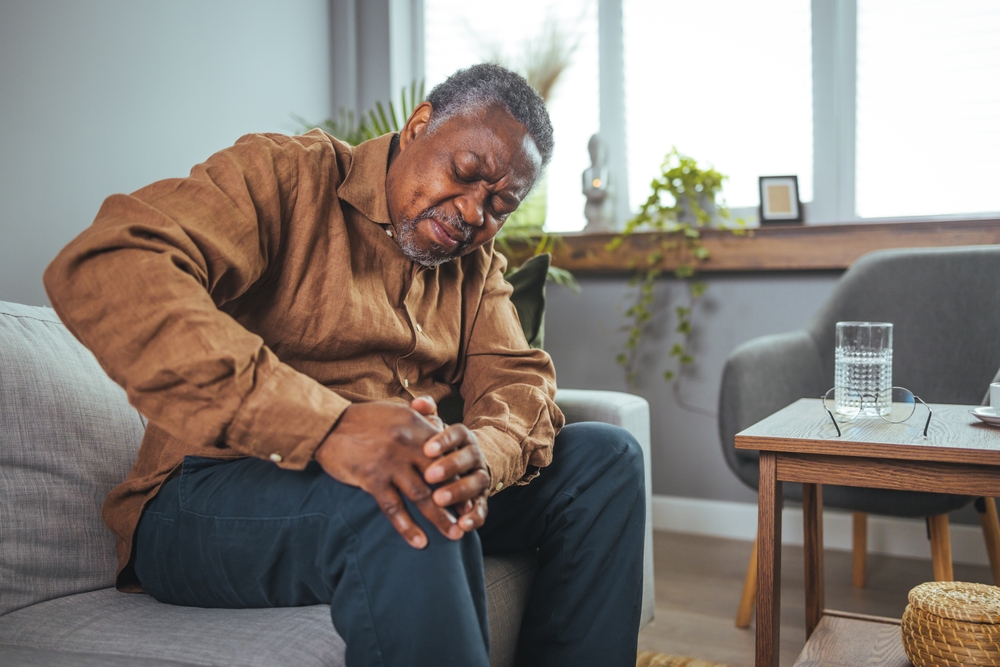 An elderly man sits on a couch, holding his knee with a pained expression.